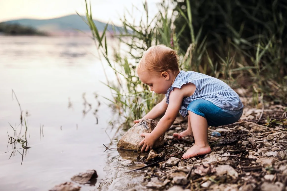 A small toddler girl playing outdoors by the river in summer.