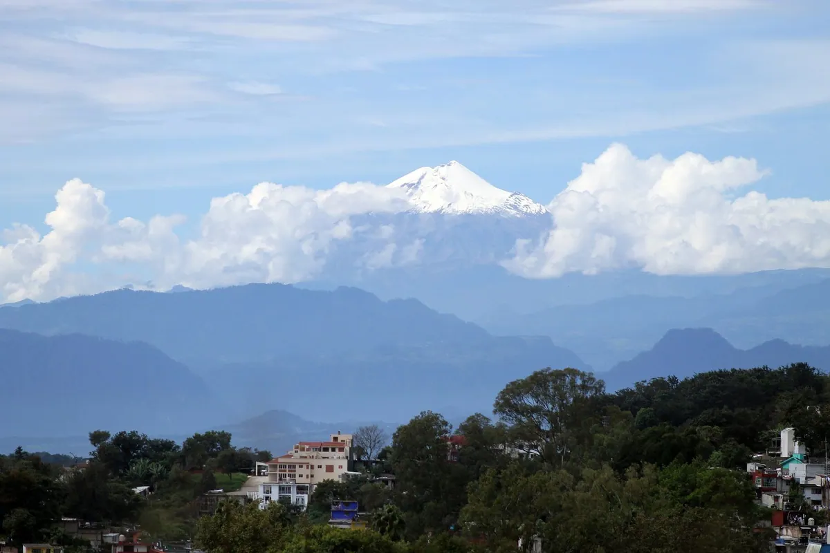 El Pico de Orizaba nevado se eleva sobre nubes, montañas y una población.